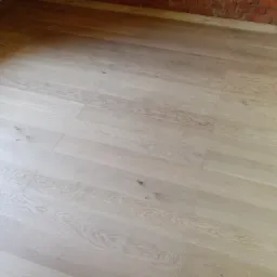 Jasna, drewniana podłoga ułożona z paneli, widoczna z góry, z fragmentem ceglanej ściany w tle, oświetlona naturalnym światłem.