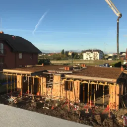 Wznoszenie ścian domu z pustaków ceramicznych; widoczna drabina oparta o ścianę, podpory stropu, oraz ramię pompy do betonu w tle.