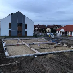 Wylane fundamenty pod budowę domu jednorodzinnego, widoczne zarys fundamentów i piasek, w tle zabudowa osiedlowa i pochmurne niebo.