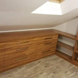 Drewniana zabudowa pod skosem z szufladami i p&oacute;łkami, wykończona w ciepłym odcieniu brązu, z metalowymi uchwytami, na podłodze panele imitujące drewno.