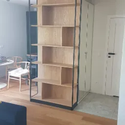 Drewniany regał z czarną metalową ramą, oddzielający przestrzeń w nowoczesnym wnętrzu, z widoczną podłogą z jasnego drewna i elementami wyposażenia.