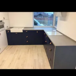 Nowoczesna kuchnia z granatowymi szafkami i jasnymi drewnianymi uchwytami, biały zlew ceramiczny pod oknem, jasna podłoga, widok na dom zza okna.