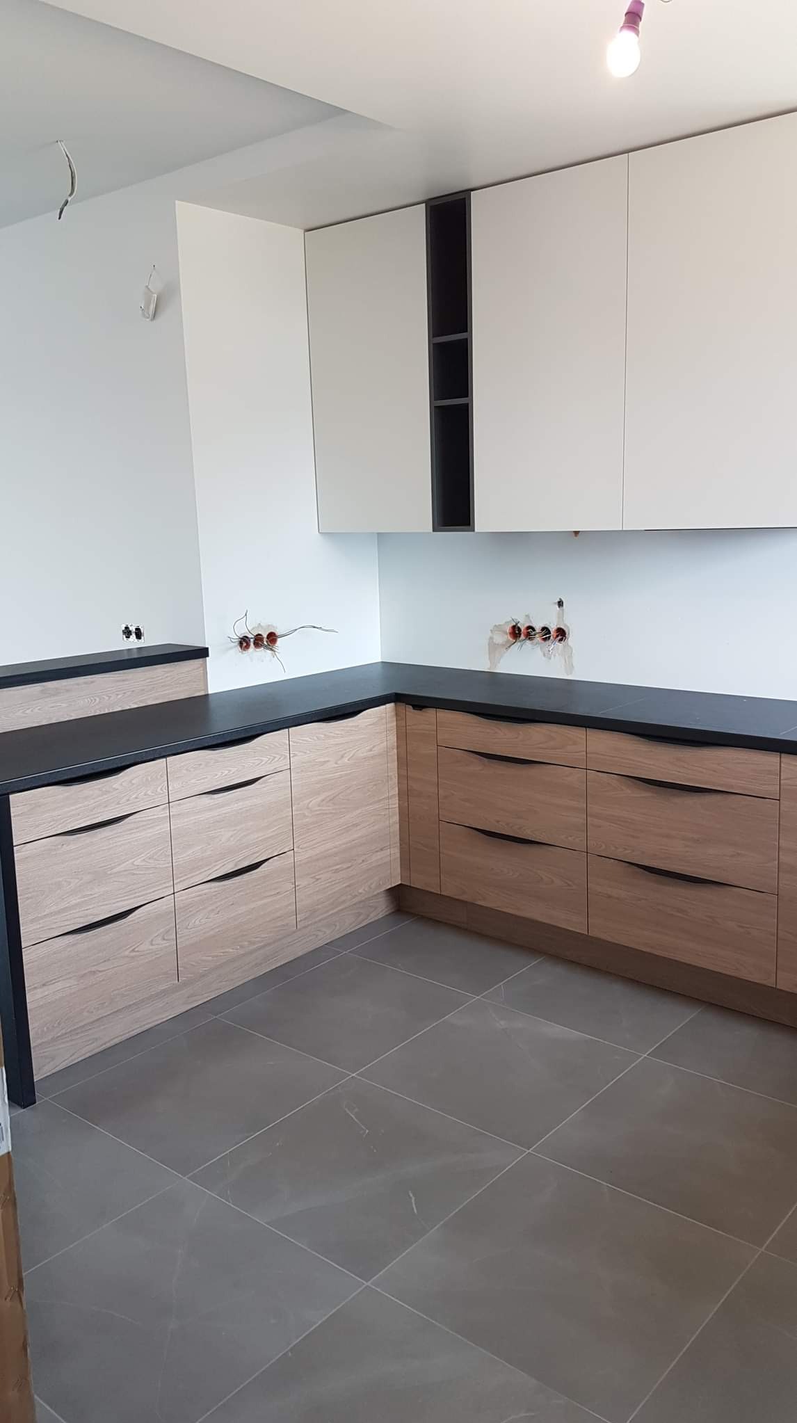 Nowoczesna, jasna kuchnia z drewnianymi frontami szuflad i czarnym blatem, widoczne niedokończone instalacje elektryczne na ścianie.