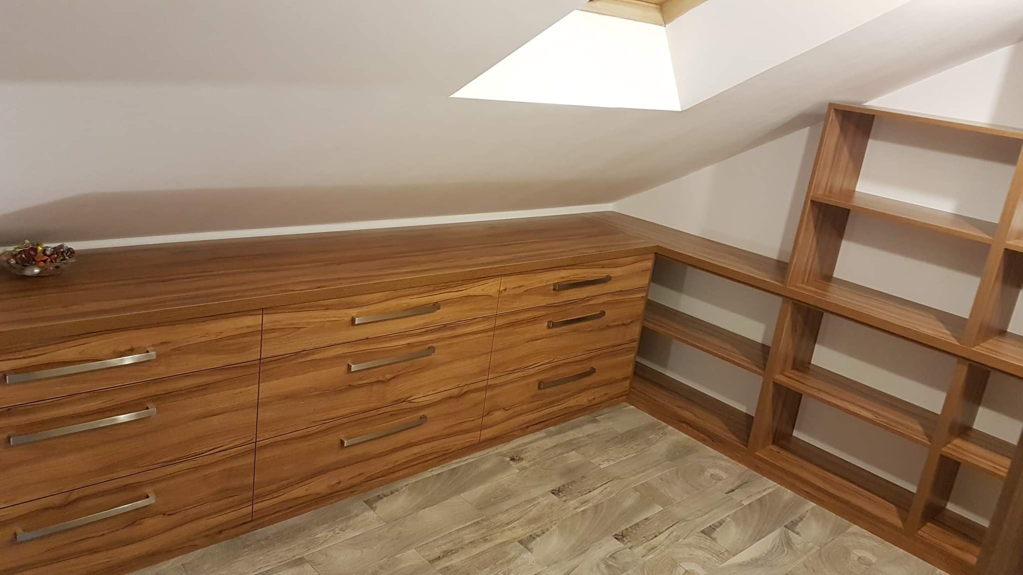 Drewniana zabudowa pod skosem z szufladami i półkami, wykończona w ciepłym odcieniu brązu, z metalowymi uchwytami, na podłodze panele imitujące drewno.