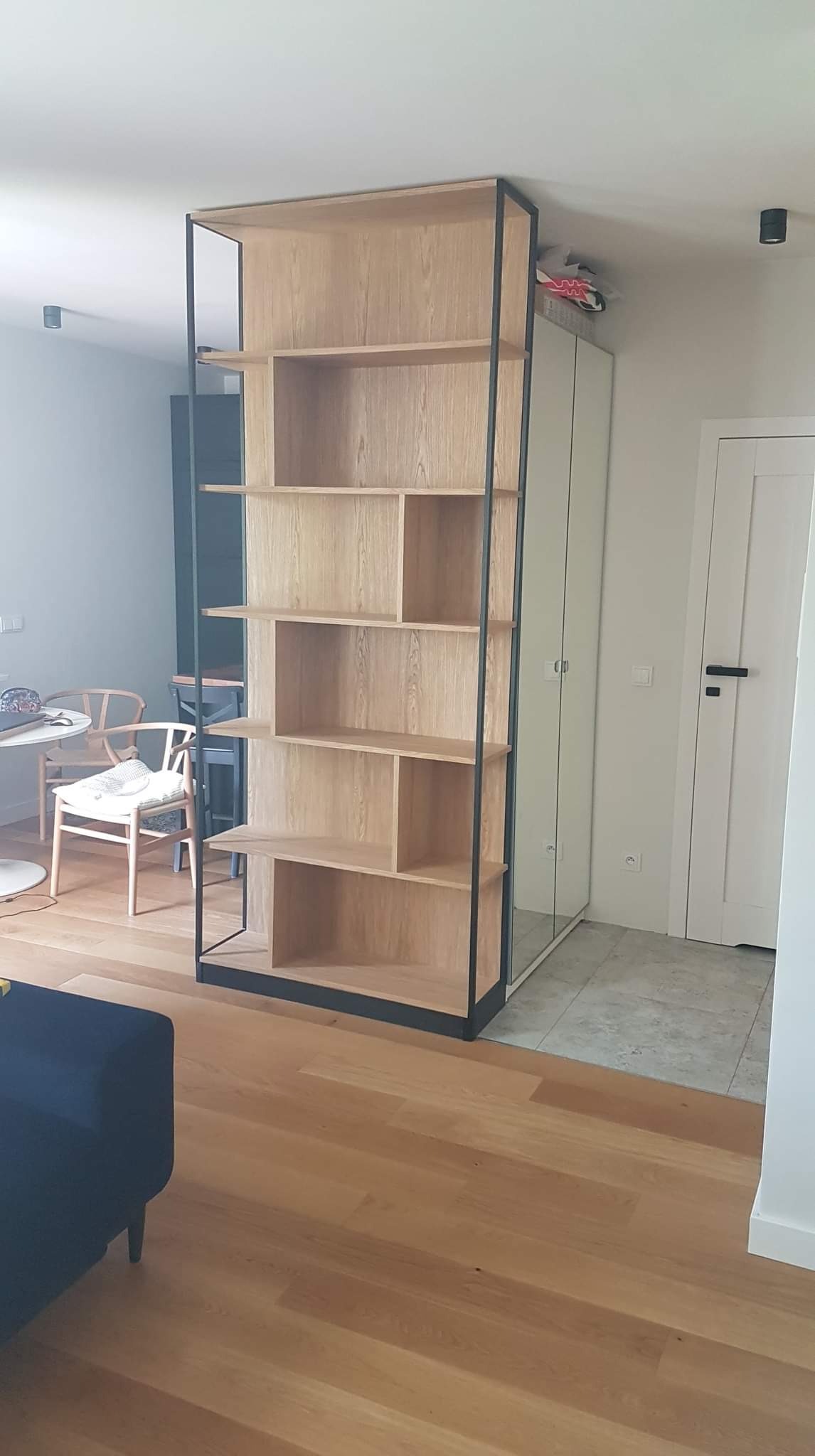 Drewniany regał z czarną metalową ramą, oddzielający przestrzeń w nowoczesnym wnętrzu, z widoczną podłogą z jasnego drewna i elementami wyposażenia.