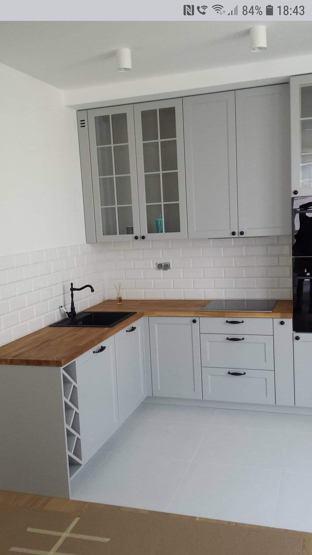 Jasnoszara kuchnia narożna z drewnianym blatem, czarnym zlewem i baterią w stylu retro, białe płytki metro na ścianie, widoczna szafka z półkami na wino.