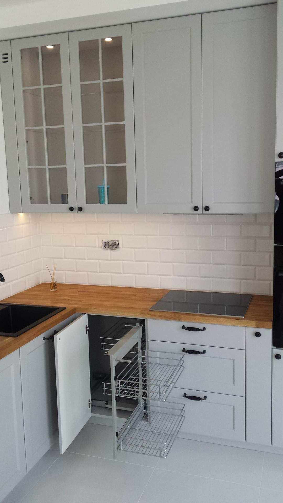 Szare szafki kuchenne z drewnianym blatem, szklanymi frontami górnych szafek i wysuwanym koszem cargo na akcesoria kuchenne.