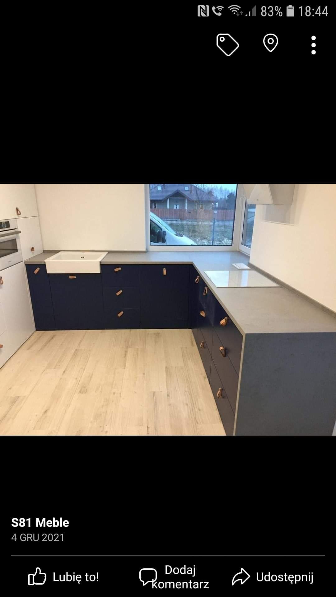 Nowoczesna kuchnia z granatowymi szafkami i jasnymi drewnianymi uchwytami, biały zlew ceramiczny pod oknem, jasna podłoga, widok na dom zza okna.