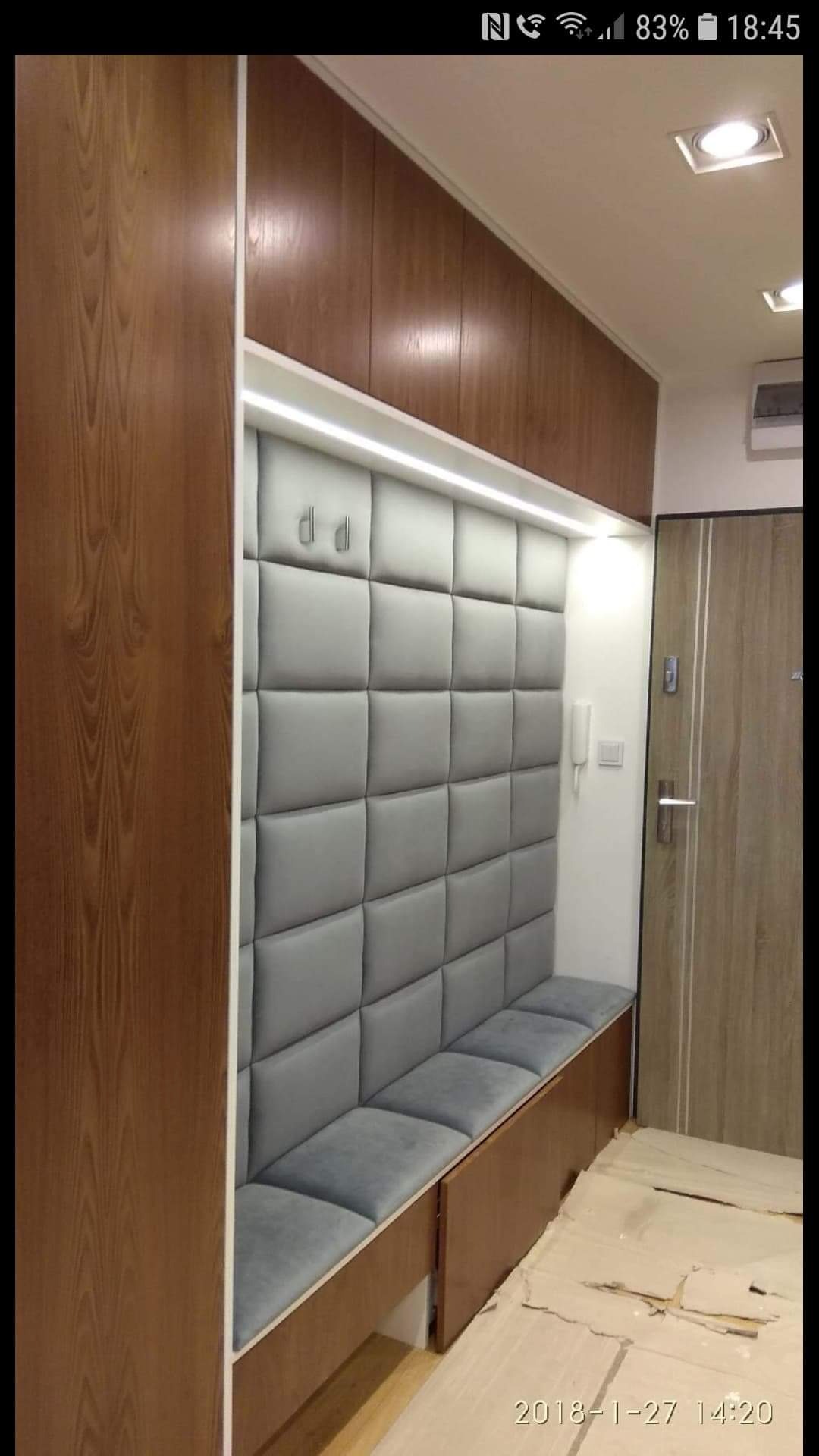 Wnęka wejściowa z siedziskiem i tapicerowanym panelem ściennym w odcieniach szarości, oświetlona listwą LED, obok drewniane szafki i drzwi wejściowe.