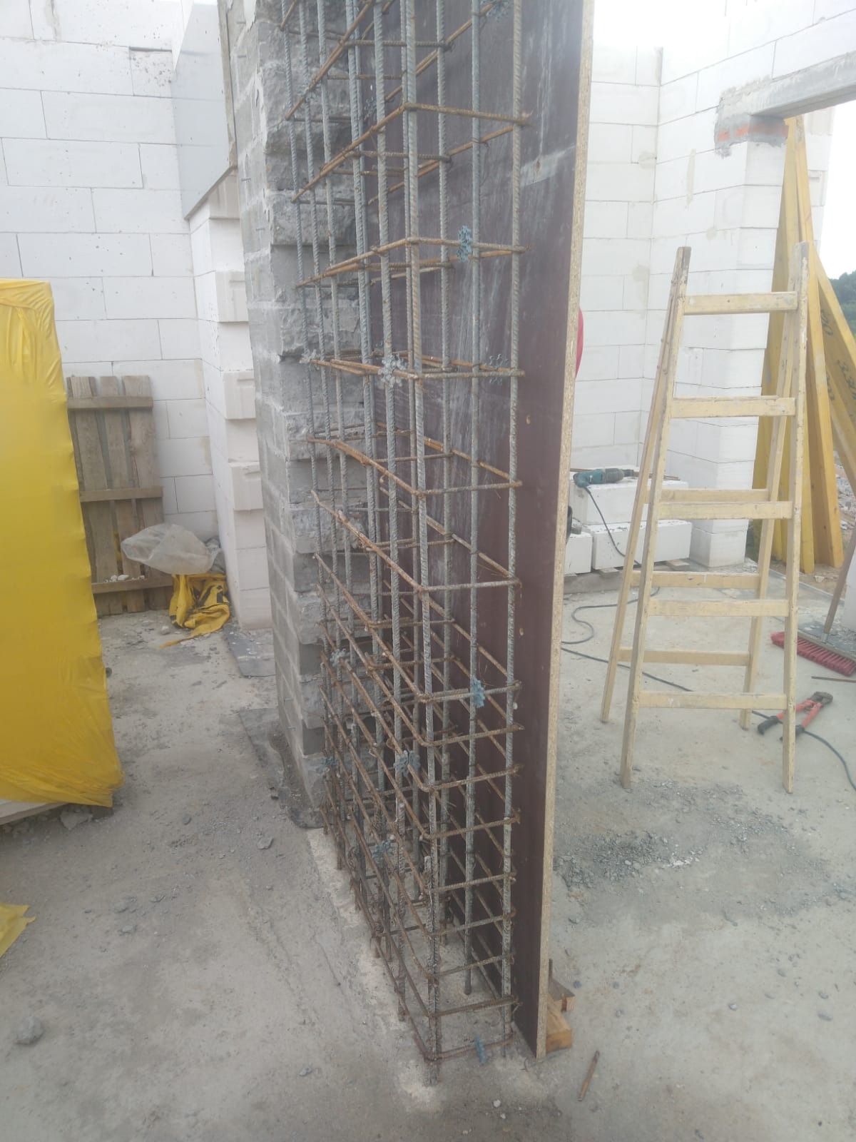 Wiązanie zbrojenia pionowego ściany z szalunkiem, widoczne białe bloczki oraz drabina w tle.