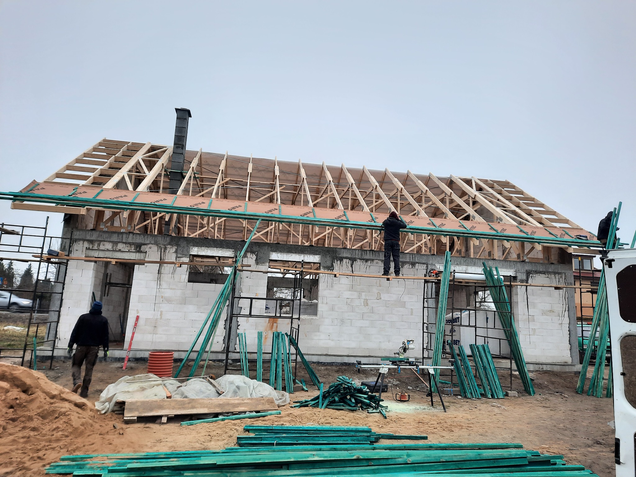 Budowa domu z białych bloczków: widoczna konstrukcja dachu z drewnianych belek, komin oraz pracownicy na rusztowaniu, na pierwszym planie stos zielonych desek.