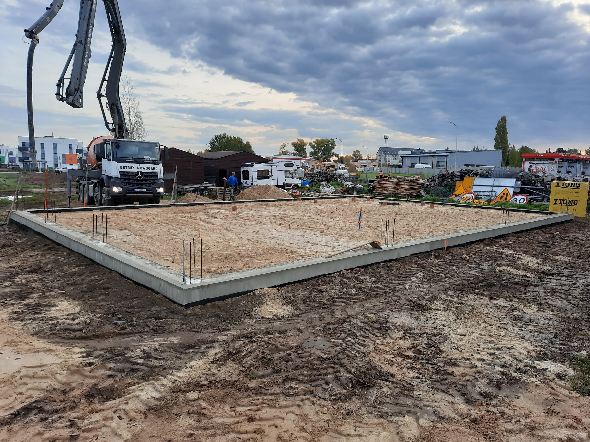 Fundament domu z wystającymi prętami zbrojeniowymi, betonowany z użyciem pompy do betonu firmy Betmix Nowogard, widoczne tablice ostrzegawcze i składowane materiały budowlane w tle.