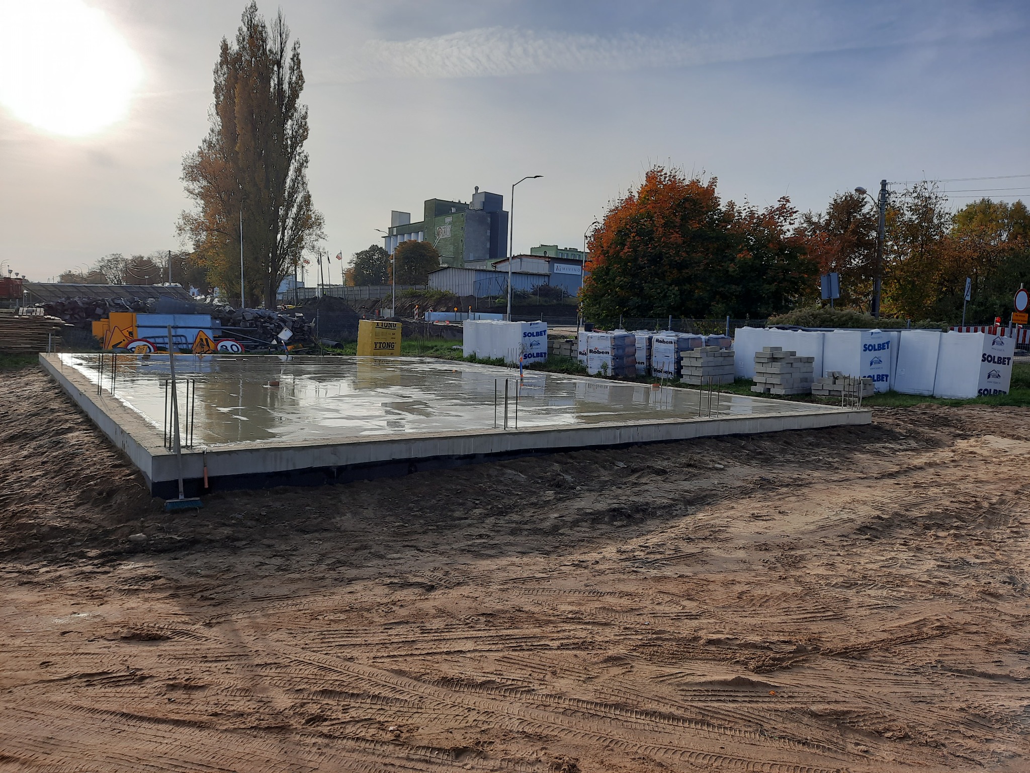 Świeżo wylana płyta fundamentowa z wystającymi prętami zbrojeniowymi, widoczne bloczki Solbet i Ytong w tle, teren budowy z widocznymi śladami opon.