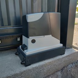 Michał Tomaszewski NetInstal - Szary napęd bramy przesuwnej Nice Robus zamontowany na betonowym fundamencie obok metalowej bramy. Widoczna listwa zębata bramy i kabel zasilający.