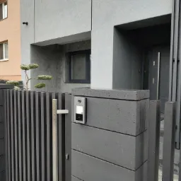 Nowoczesna, szara brama z pionowych sztachet i elementami z betonu architektonicznego, z zamontowanym wideodomofonem i ozdobnym drzewkiem bonsai na tle domu w podobnej kolorystyce.