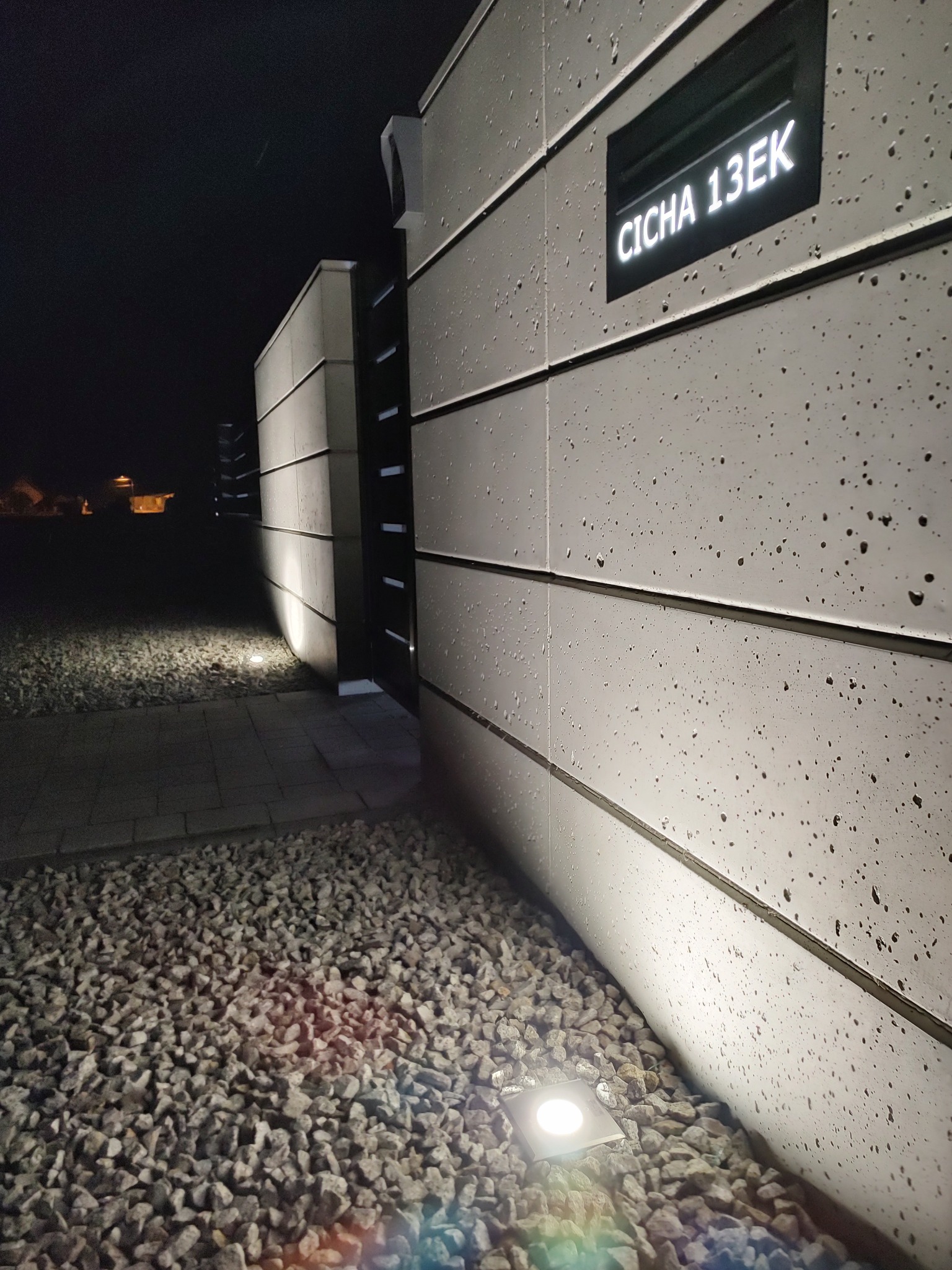 Nowoczesne oświetlenie posesji nocą: podświetlony numer domu 'CICHA 13EK' i lampa gruntowa na żwirowym podłożu, widoczne krople deszczu na elewacji.