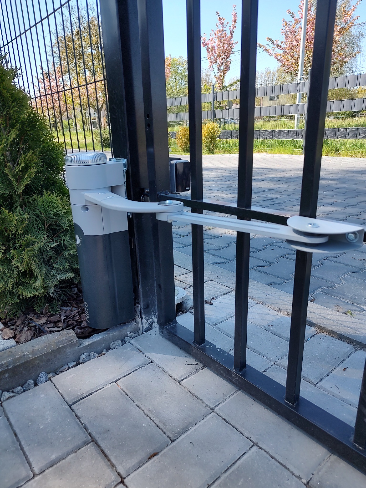 Zamontowany napęd bramy skrzydłowej Nice na tle metalowego ogrodzenia i kostki brukowej, z widocznym krzewem iglastym po lewej stronie.