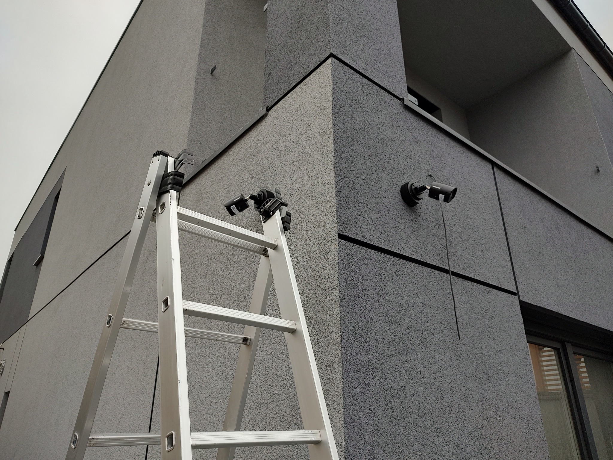 Aluminiowa drabina oparta o szarą elewację nowoczesnego domu, widoczne dwie zainstalowane kamery monitoringu na ścianie.