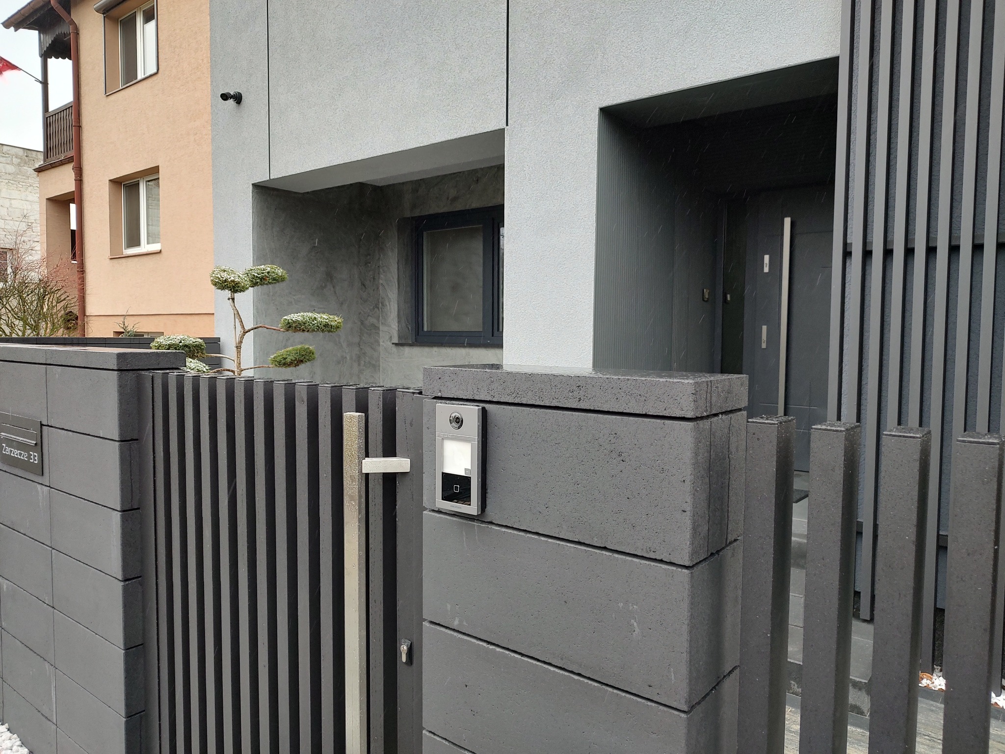 Nowoczesna, szara brama z pionowych sztachet i elementami z betonu architektonicznego, z zamontowanym wideodomofonem i ozdobnym drzewkiem bonsai na tle domu w podobnej kolorystyce.