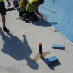 Układanie jasnoniebieskich płytek na posadzce, widoczni pracownicy w odblaskowych kamizelkach i szarych szortach, jeden w ruchu, rzucający cień na jasną powierzchnię, obok ułożone płytki i czarne...