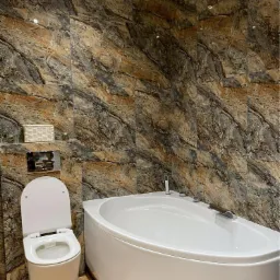 Biała, narożna wanna i podwieszana toaleta w łazience wykończonej płytkami imitującymi marmur w odcieniach brązu i szarości, z akcentami dekoracyjnymi na półce nad spłuczką.