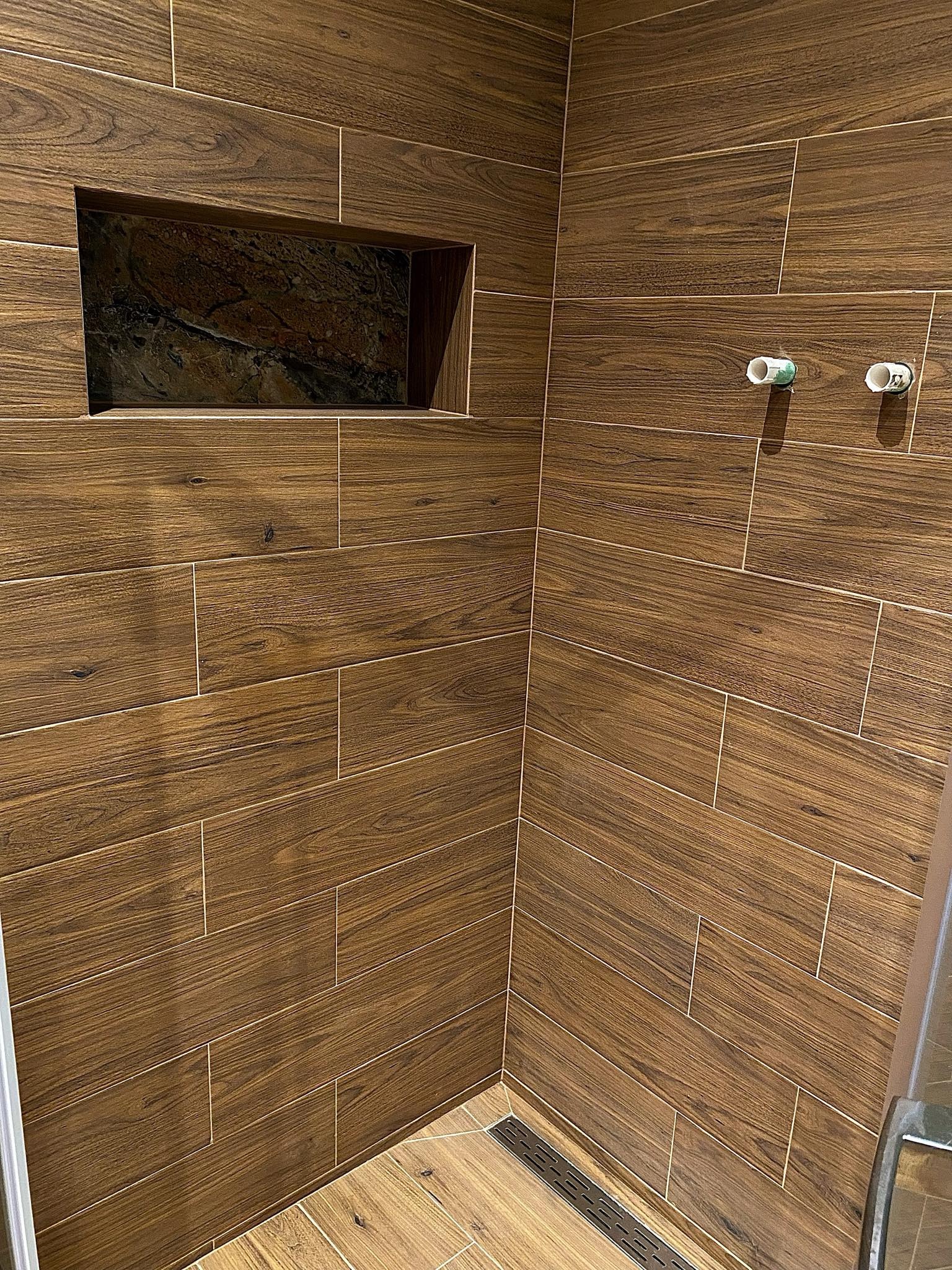 Wykończona kabina prysznicowa z brązowymi płytkami imitującymi drewno ułożonymi poziomo, w ścianie wnęka wyłożona kamieniem, widoczne dwa wystające rury z zaślepkami.