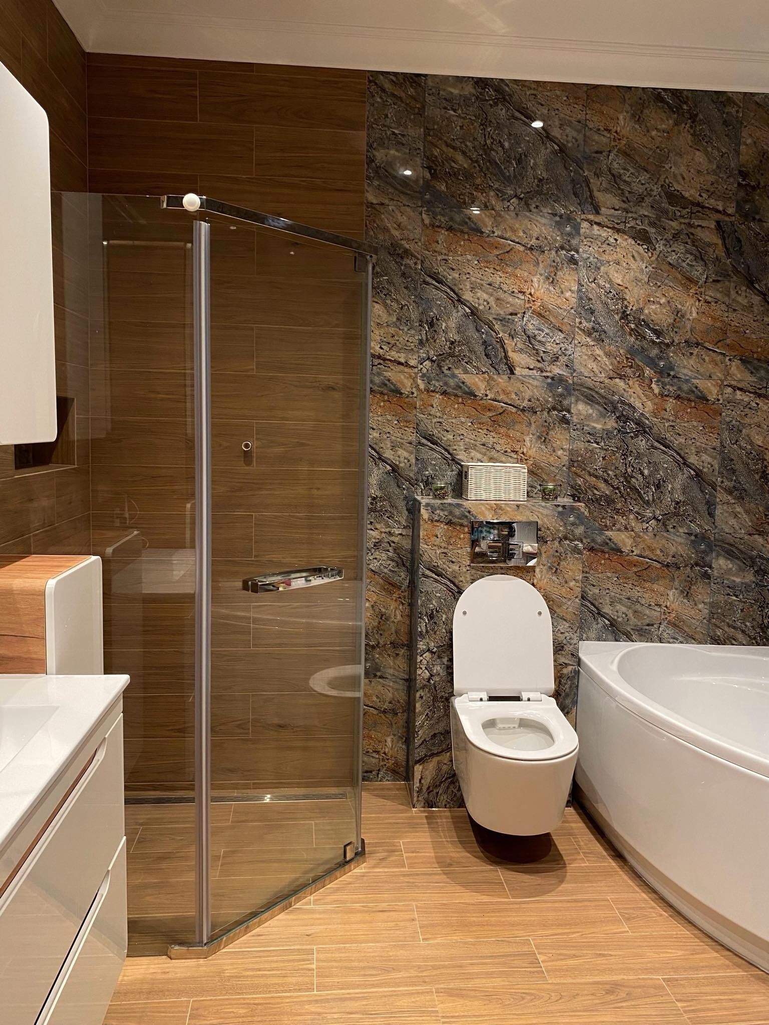 Nowoczesna łazienka z kabiną prysznicową ze szkła, toaletą typu Geberit, wanną narożną i płytkami imitującymi drewno oraz marmur.