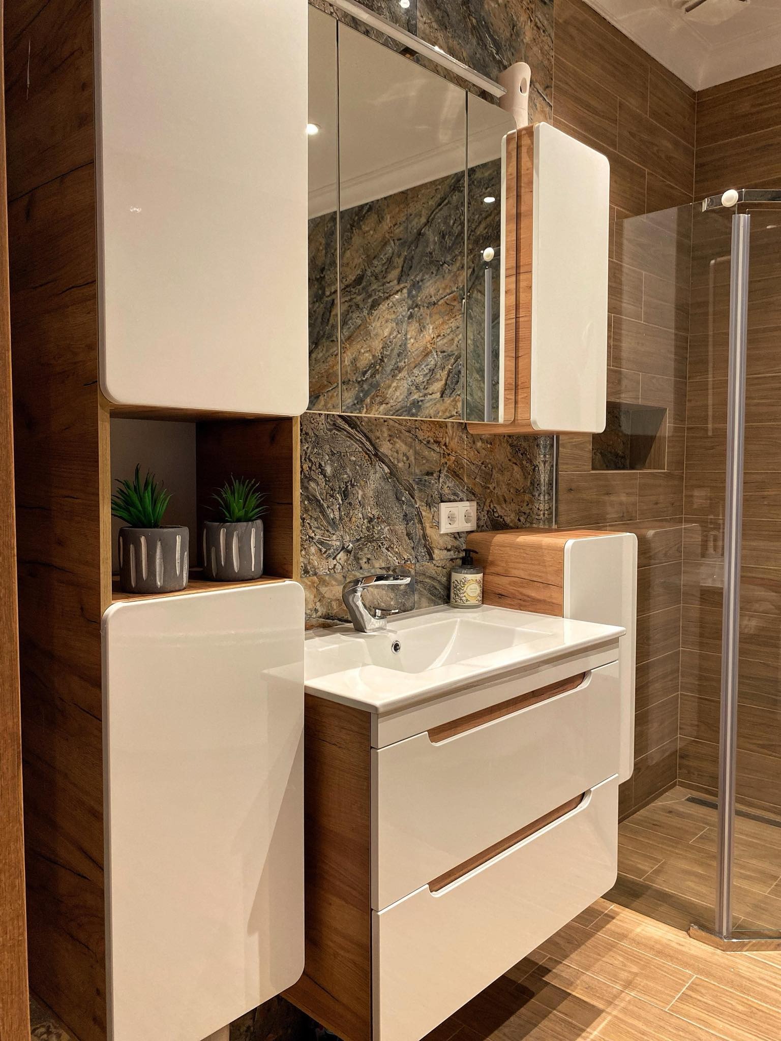 Nowoczesna łazienka z drewnopodobną glazurą, białą szafką umywalkową z drewnianymi akcentami, lustrem w szafce i kabiną prysznicową w tle.