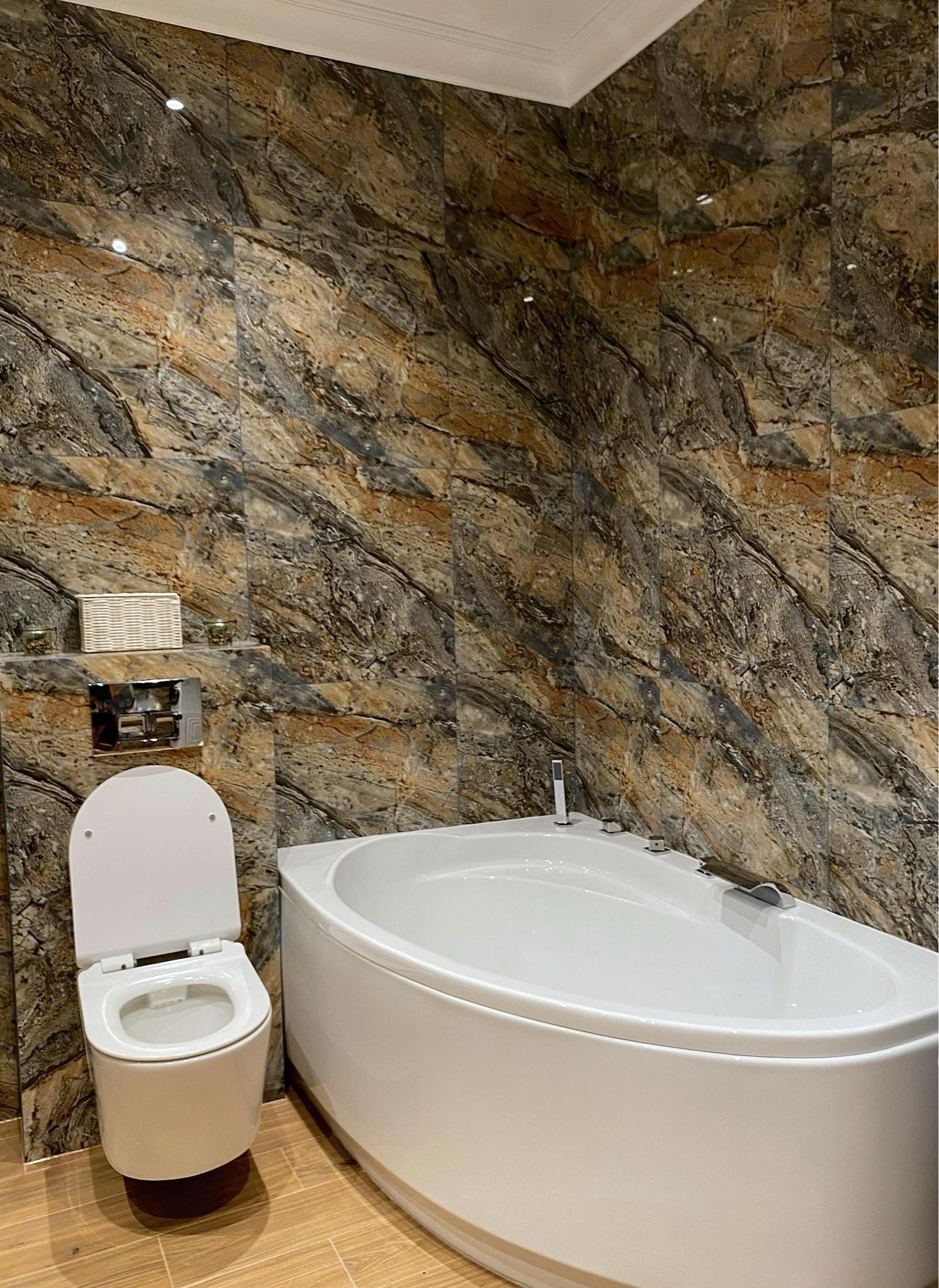 Biała, narożna wanna i podwieszana toaleta w łazience wykończonej płytkami imitującymi marmur w odcieniach brązu i szarości, z akcentami dekoracyjnymi na półce nad spłuczką.