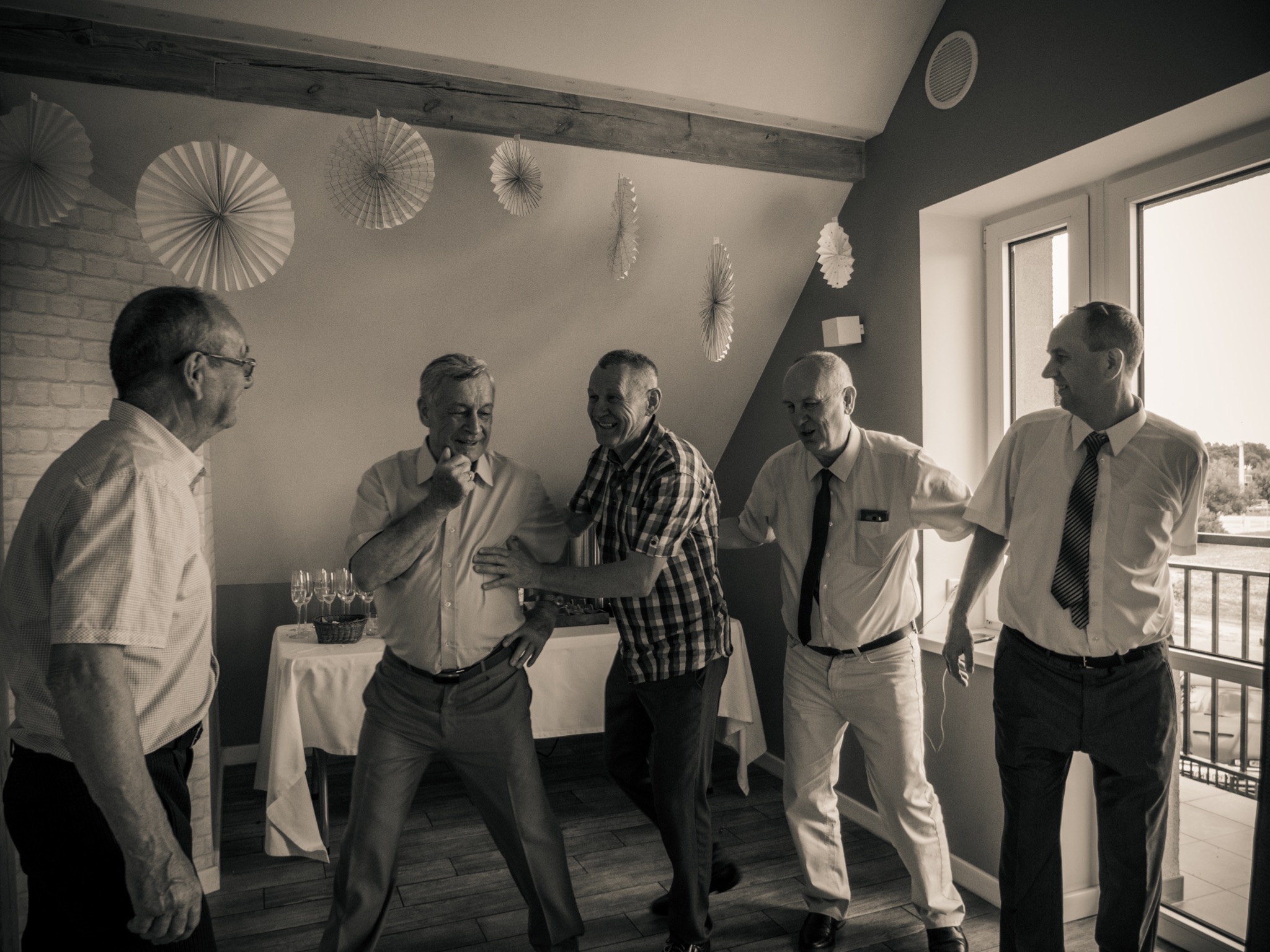 Czarno-białe zdjęcie przedstawia pięciu mężczyzn w średnim wieku, prawdopodobnie uczestniczących w zabawie lub tańcu podczas wydarzenia. W tle widoczne dekoracje papierowe i stół z kieliszkami.