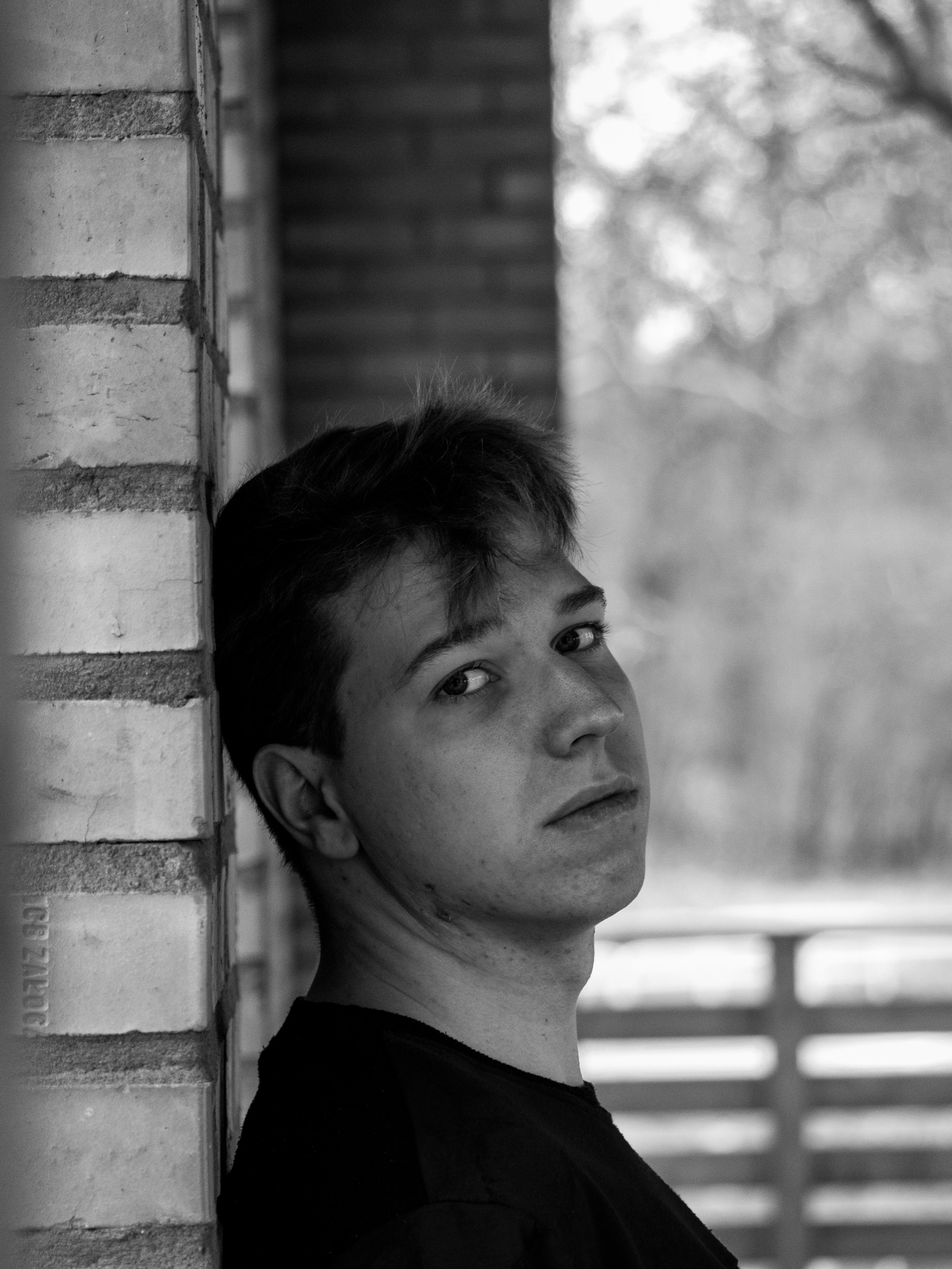 Czarno-biały portret młodego mężczyzny opierającego się o ceglaną ścianę, zamyślony wyraz twarzy, widoczne pojedyncze cegły i rozmyte tło z elementami roślinności.