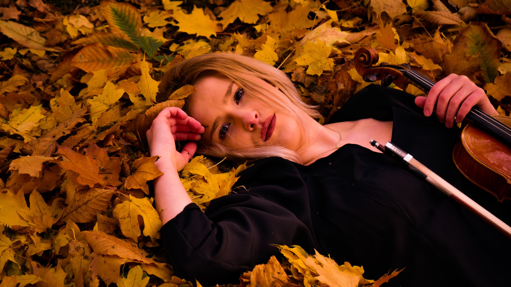 Blondwłosa kobieta w czarnej sukience leży na ziemi pokrytej żółtymi liśćmi, trzymając skrzypce i smyczek.