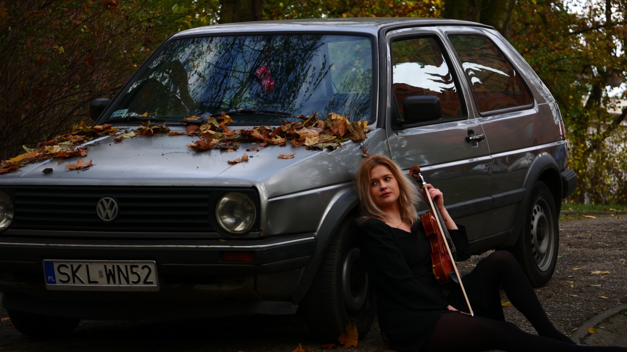 Młoda kobieta w czarnym ubraniu siedzi oparta o srebrny samochód Volkswagen Golf, trzymając skrzypce; jesienne liście na masce auta.