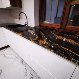 Kamieniarstwo.pl - Czarny blat kuchenny z unikalnym złotym użyleniem, wkomponowany w białą zabudowę kuchenną przy oknie z drewnianą ramą. Widoczny czarny zlew i bateria. Na podłodze białe płytki imitujące marmur.