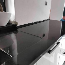 Kamieniarstwo.pl - Błyszczący, czarny blat z kamienia z wyciętym otworem, zamontowany na białych szafkach z podłużnymi, czarnymi uchwytami, obok doniczka z bonsai, widoczny fragment ściany z włącznikiem.