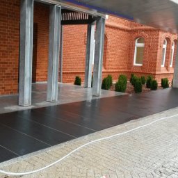 Kamieniarstwo.pl - Nowoczesny taras z ciemnych płyt kamiennych przy budynku z czerwonej cegły, widoczne metalowe słupy i fragment węża ogrodowego na jasnej kostce brukowej.