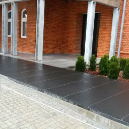 Nowoczesny taras z ciemnych płyt kamiennych, z widoczną kostką brukową i ceglaną elewacją budynku w tle.