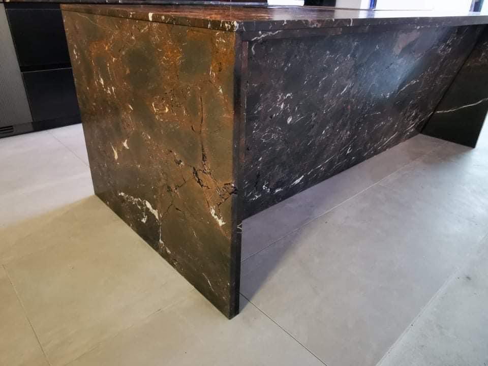 Wyspa kuchenna z kamiennym blatem o ciemnej kolorystyce z wyraźnym, nieregularnym wzorem żyłkowania w odcieniach bieli i brązu, ustawiona na jasnej, matowej podłodze.