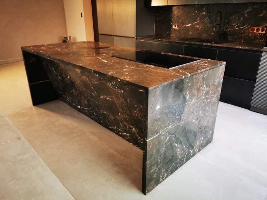 Nowoczesna wyspa kuchenna z ciemnego kamienia z wyraźnym żyłkowaniem, dopasowana do ciemnej zabudowy kuchennej w tle, oświetlenie górne.