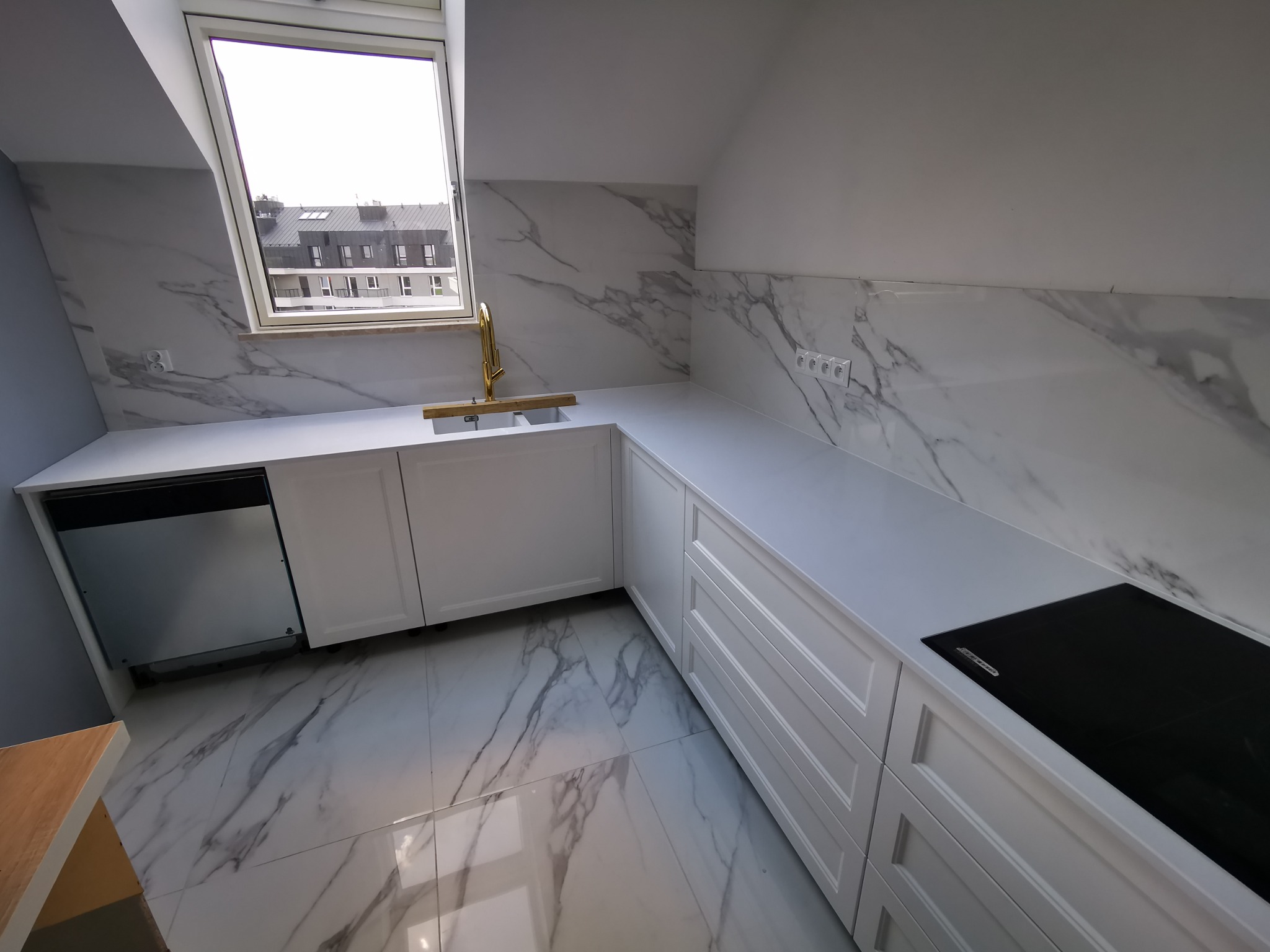 Biała kuchnia z marmurowym wzorem na ścianie i podłodze, ze złotą armaturą przy zlewie pod oknem dachowym.