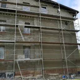 Dwupiętrowy budynek w trakcie prac elewacyjnych, z widocznym rusztowaniem, częściowo otynkowaną ścianą i zabezpieczonymi oknami folią. Na dole widoczne pozostałości po pracach i puste wiaderka.
