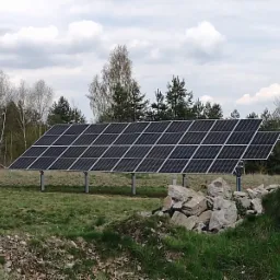 Dwa rzędy paneli fotowoltaicznych zamontowane na metalowych podporach na otwartej przestrzeni, z stertą szarych kamieni na pierwszym planie i drzewami w tle pod pochmurnym niebem.