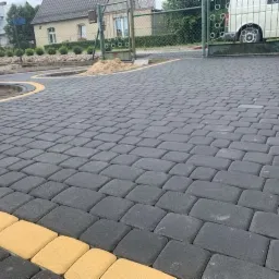 Ułożona, szara kostka brukowa z żółtym obrzeżem, prowadząca do metalowej bramy wjazdowej i domu z jasną elewacją w tle.