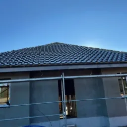 Dom w trakcie prac elewacyjnych z widocznym rusztowaniem, szarą elewacją i nowym, ciemnym dachem z dach&oacute;wki ceramicznej.