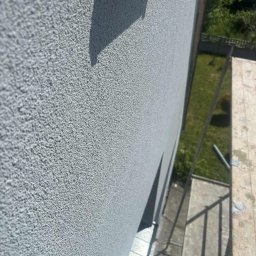 Kacper Skorb Usługi Budowlane - Szara, ziarnista elewacja budynku z bliska, widziana z rusztowania. Świeżo nałożony tynk strukturalny w neutralnym odcieniu szarości.