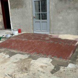 Kacper Skorb Usługi Budowlane - Świeżo ułożona, czerwona kostka brukowa przed wejściem do budynku. Widoczne ślady piasku i narzędzia. Szara elewacja i drzwi wejściowe.
