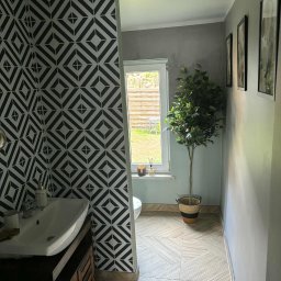 Kacper Skorb Usługi Budowlane - Wzorzysta tapeta w romby w łazience w Iłowej. Umywalka z szafką, okno z widokiem na zewnątrz, podłoga w jasne pasy. Dekoracyjne drzewko w rogu.