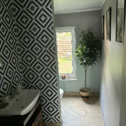 Wzorzysta tapeta w romby w łazience w Iłowej. Umywalka z szafką, okno z widokiem na zewnątrz, podłoga w jasne pasy. Dekoracyjne drzewko w rogu.