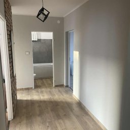 KS Budownictwo - Jasne panele podłogowe w korytarzu z szarą ścianą i ozdobną cegłą. W tle widoczna łazienka z wanną. Geometryczna lampa sufitowa dodaje nowoczesnego akcentu.