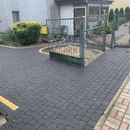 Kacper Skorb Usługi Budowlane - Ułożona szara kostka brukowa z żółtymi akcentami przy posesji z zielonym metalowym ogrodzeniem i bramą. Widoczny fragment budynku i roślinność.